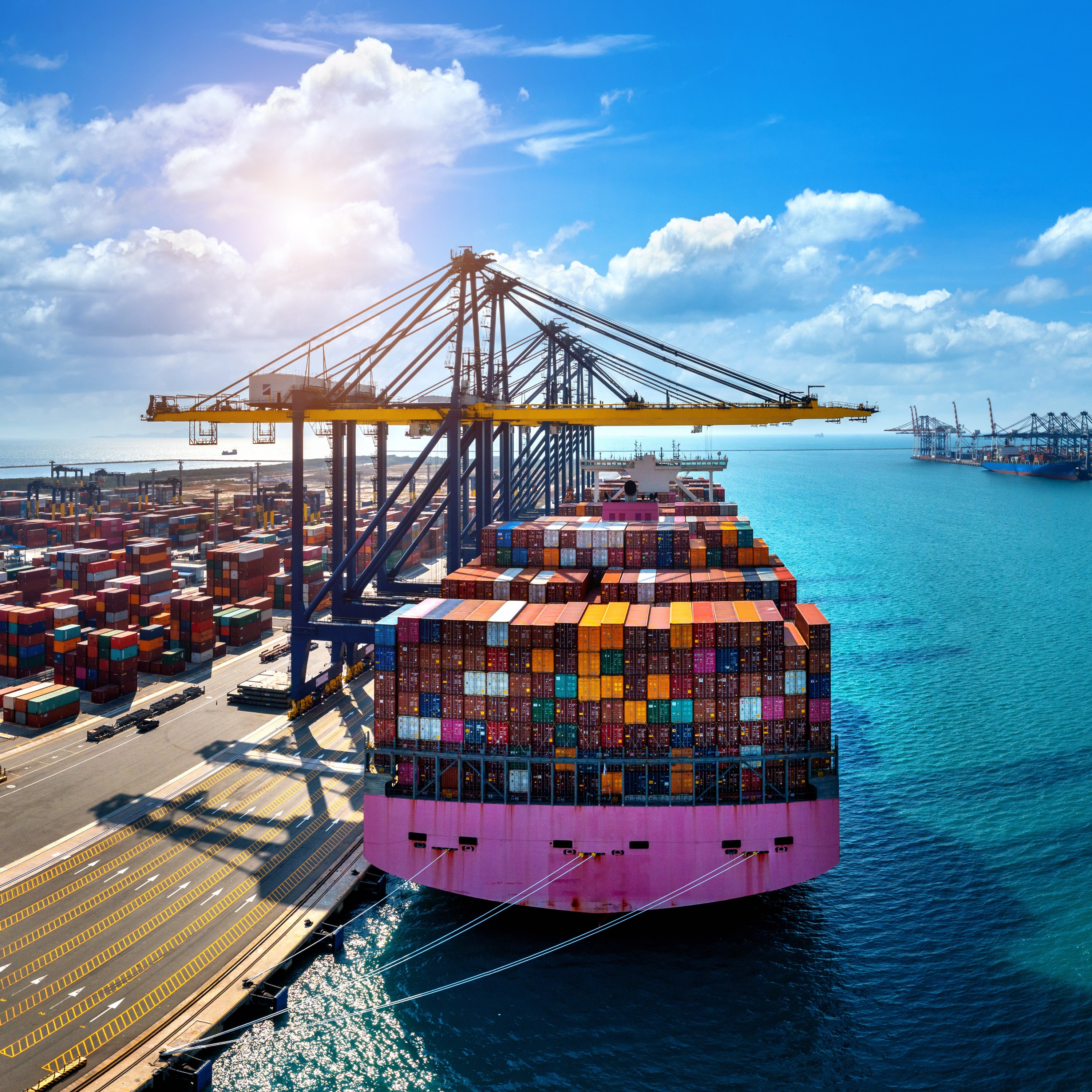Large cargo ship loaded with colorful shipping containers docked at a busy port, with cranes unloading goods and additional containers stacked along the waterfront